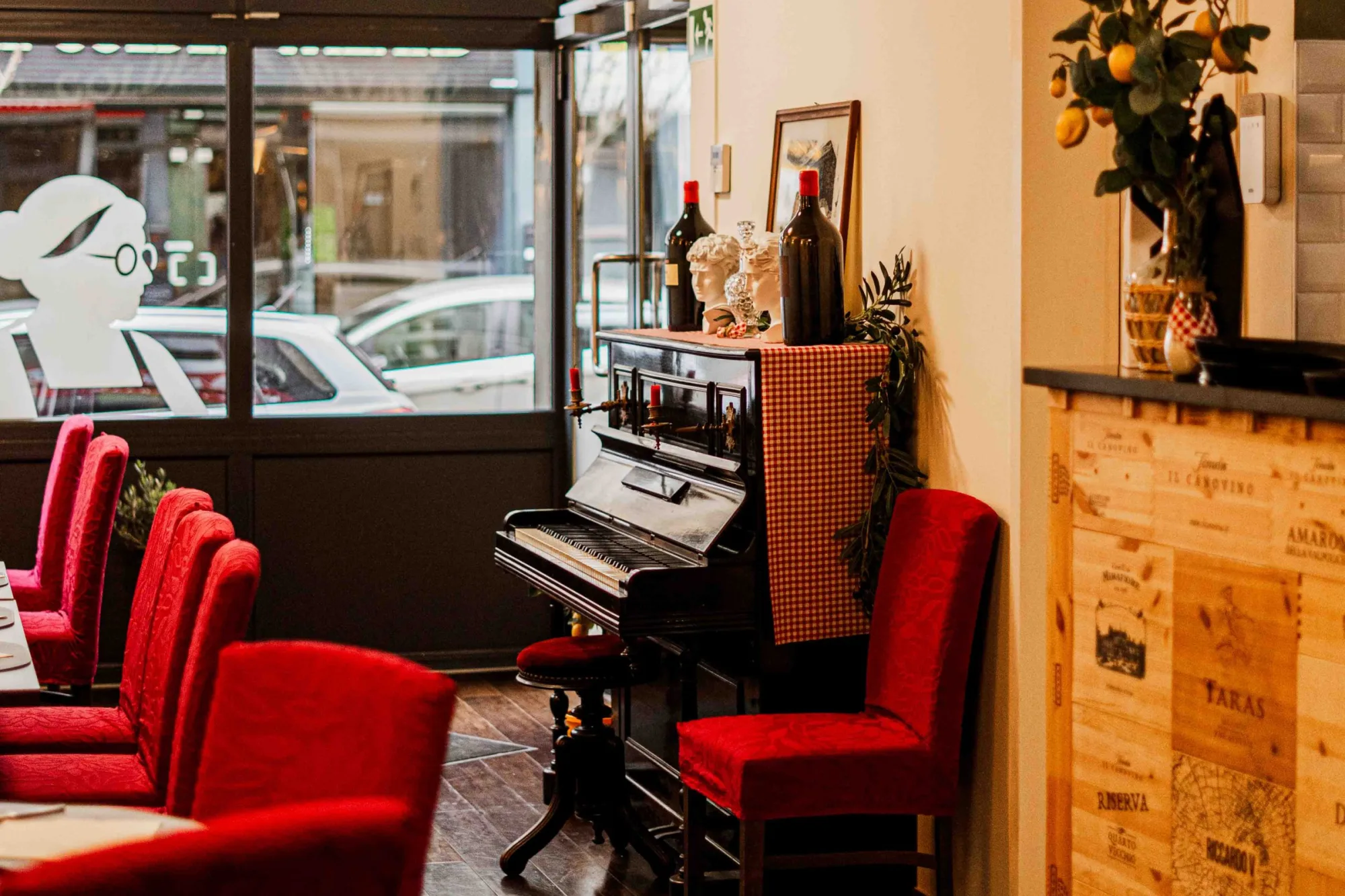 Coin salle du restaurant près de la vitrine, piano noir, décor italien et fauteuils rouges disposés autour des tables