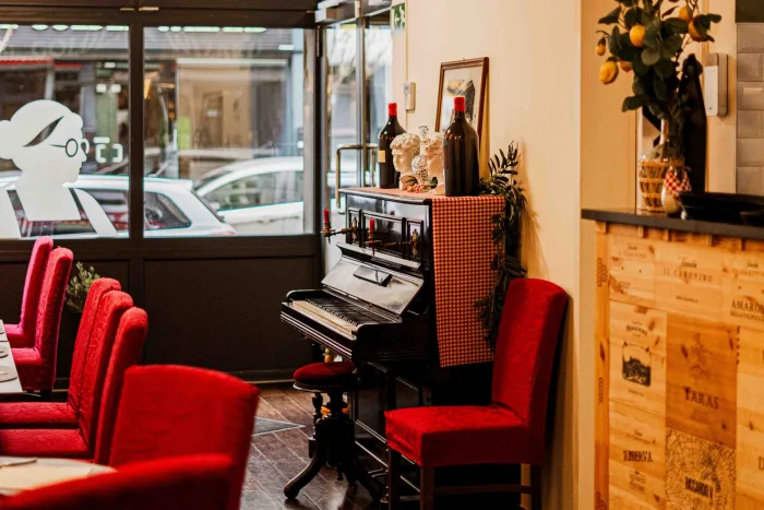 Coin salle du restaurant près de la vitrine, piano noir, décor italien et fauteuils rouges disposés autour des tables