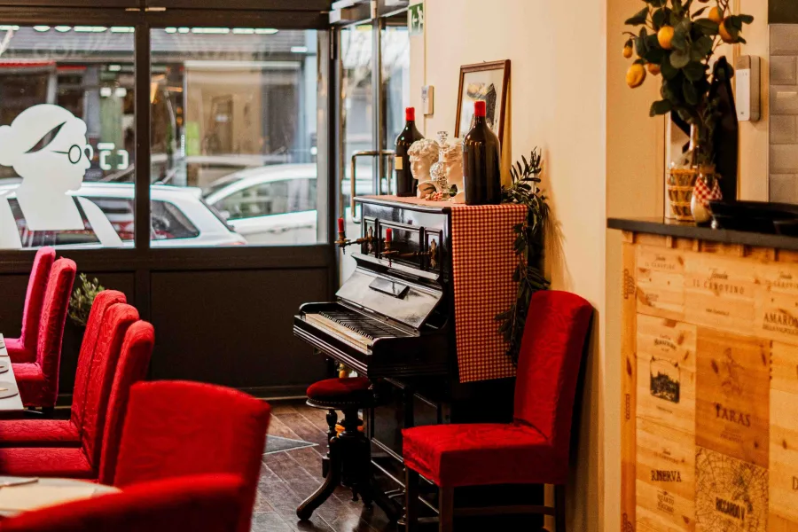 Coin salle du restaurant près de la vitrine, piano noir, décor italien et fauteuils rouges disposés autour des tables