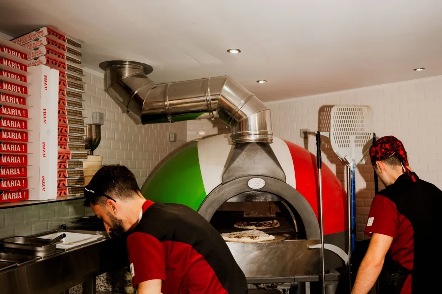 Deux pizzaiolos en tenue rouge et noire travaillent devant le four à bois, boîtes empilées derrière eux